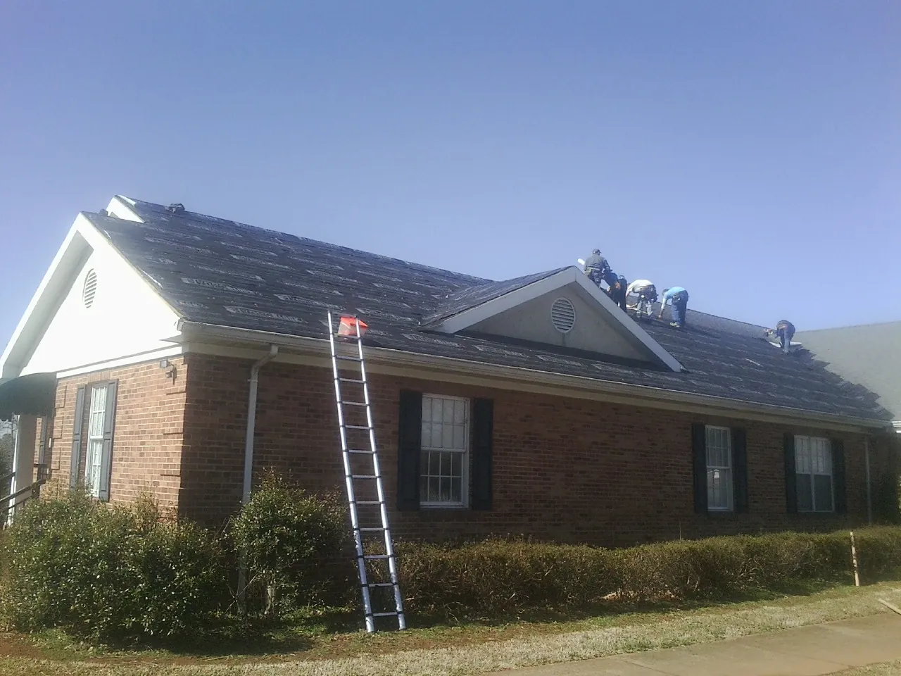 Crew tearing shingles off a brick ranch roof with a ladder set — Metal Roof Repair work by our crew in Sergeant Bluff