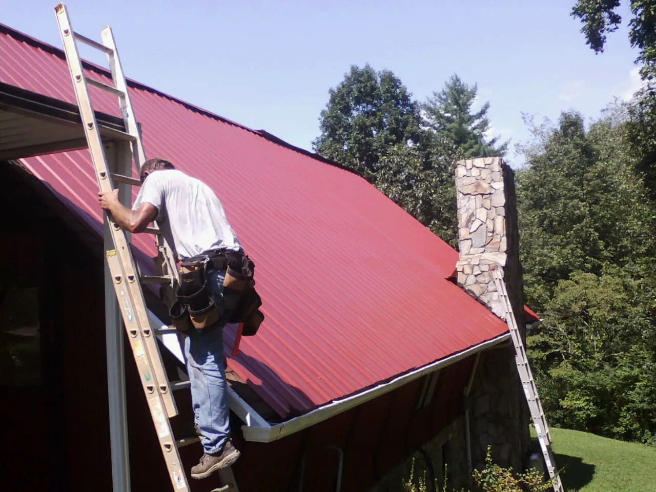 Roofer on a ladder beside a red metal roof and stone chimney — Soffit & Fascia Repair by Roofing Brothers in Sergeant Bluff